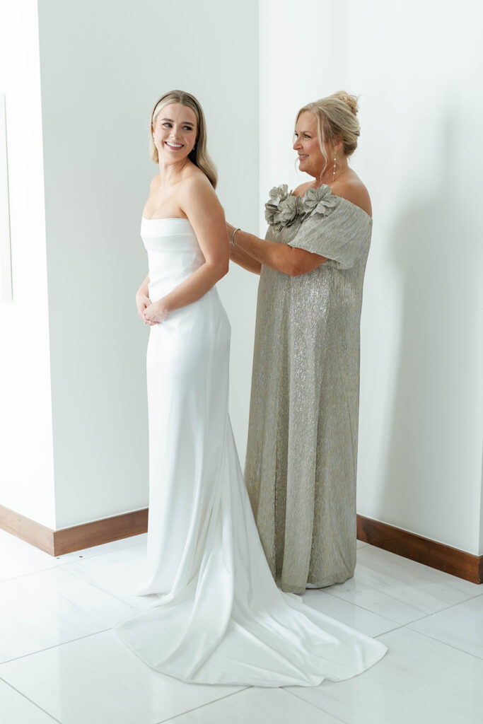 The bride putting on her wedding dress with the help of her mother and bridesmaids, while her veil is carefully adjusted and the finishing touches are added, with excitement and emotion filling the air.