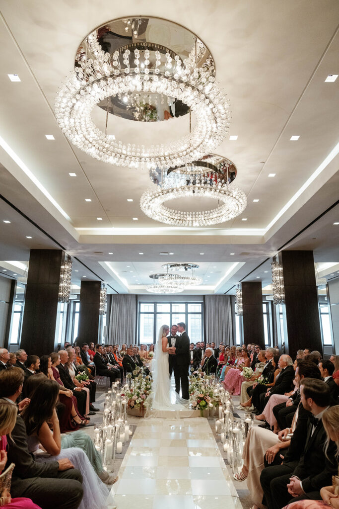 The bride walking down the aisle toward her groom, her dress flowing behind her, while the guests stand in awe, with beautiful flowers and greenery framing the serene ceremony setting.