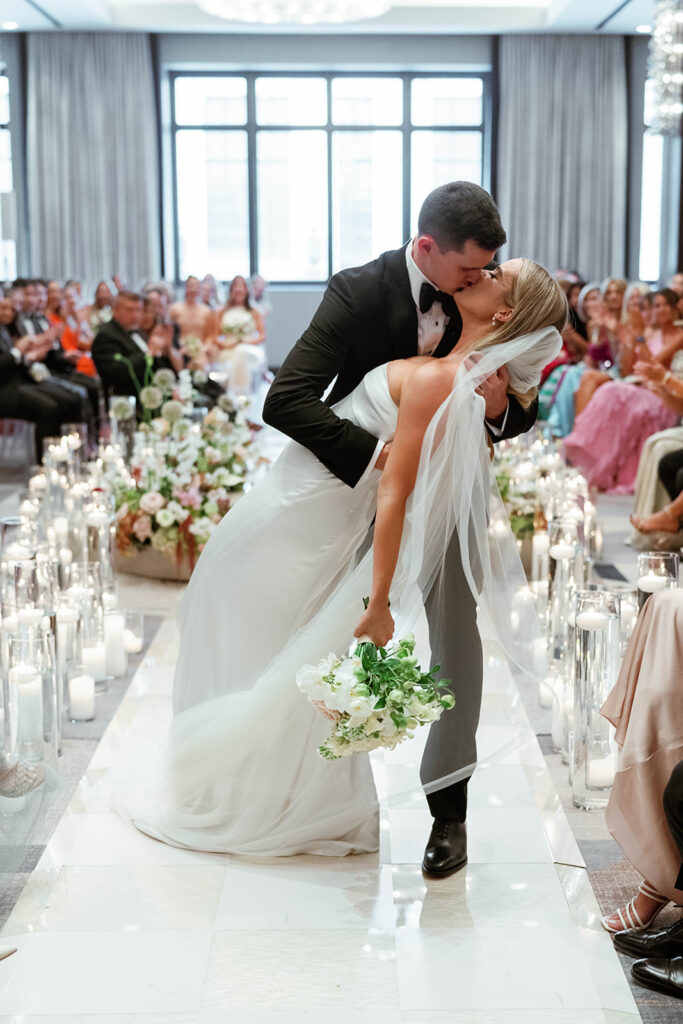 A couple sharing their first kiss as newlyweds on an elevated platform surrounded by lush floral arrangements in soft pastel hues. The venue features floor-to-ceiling windows and sparkling crystal chandeliers overhead.