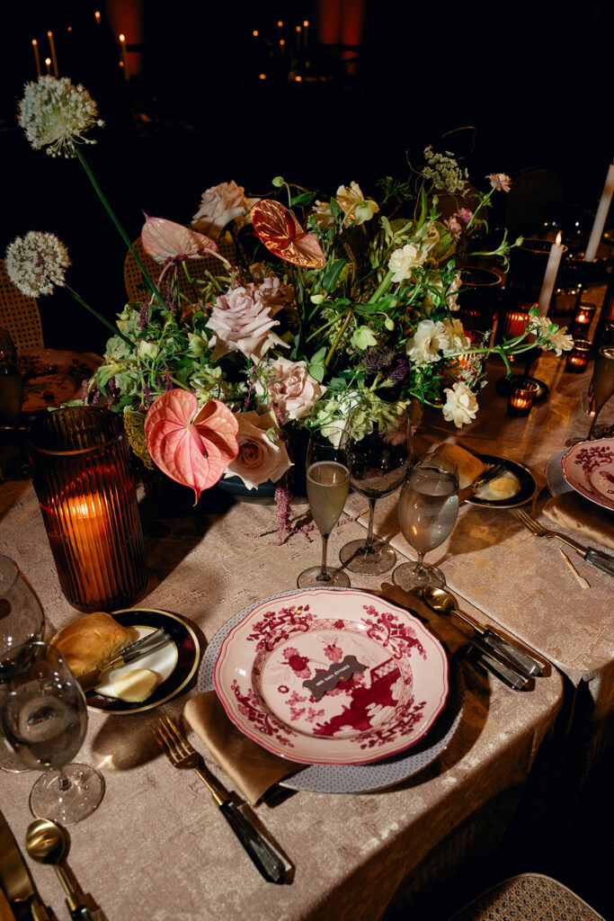 A close-up of a beautifully set reception table featuring intricate red-and-white patterned china, lush floral centerpieces with anthuriums and roses, and glowing candlelight creating an intimate atmosphere.