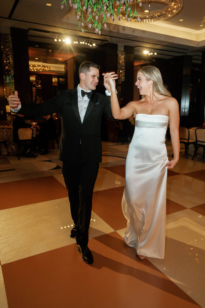 A couple dancing in an elegant ballroom with warm lighting and luxurious decor. The groom is dressed in a classic black tuxedo, while the bride wears a sleek strapless satin gown with a flowing silhouette.