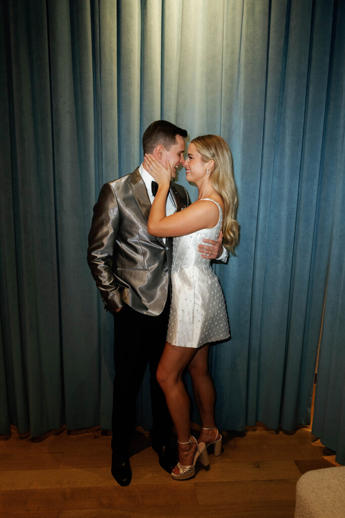 A couple posing in a modern, reflective space with city lights visible through the windows. The groom wears a silver tuxedo jacket, and the bride is dressed in a chic short white dress with textured details, paired with platform heels.