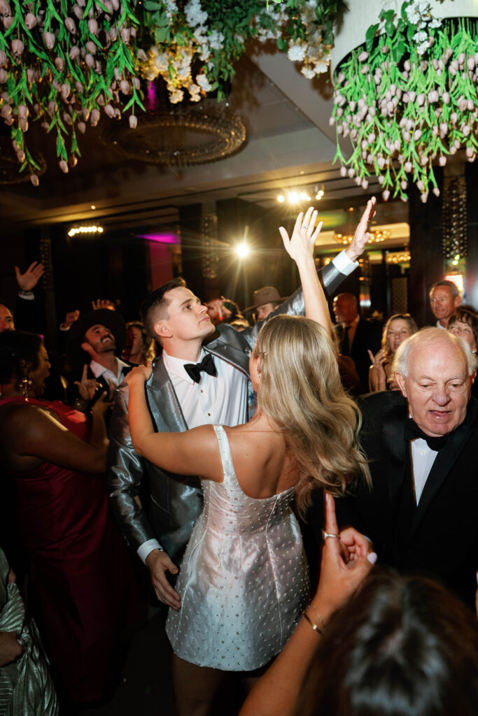 The wedding reception in full swing, with guests dancing joyfully under elegant string lights, while the bride and groom share their first dance as a married couple, surrounded by loved ones.