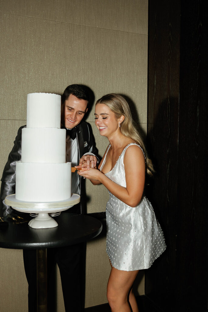 Guests enjoy the lively reception, with the bride and groom cutting their wedding cake, while friends and family cheer and clap in the background, surrounded by sparkling decorations and vibrant colors.