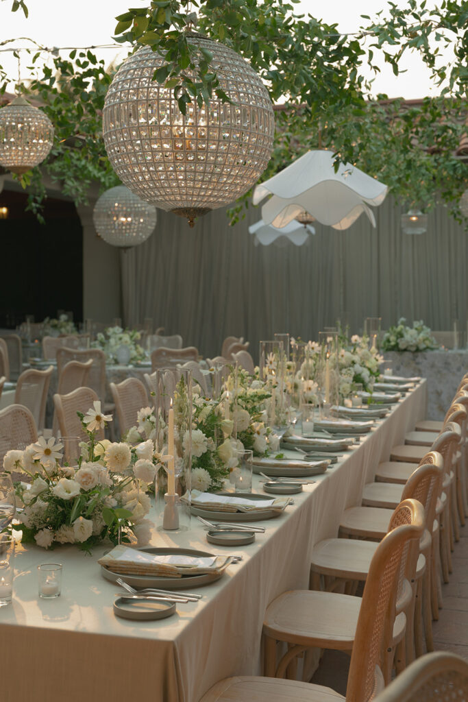 Reception tables at The Ritz-Carlton Bacara with soft white florals, candlelight, and crystal chandeliers glowing at sunset.