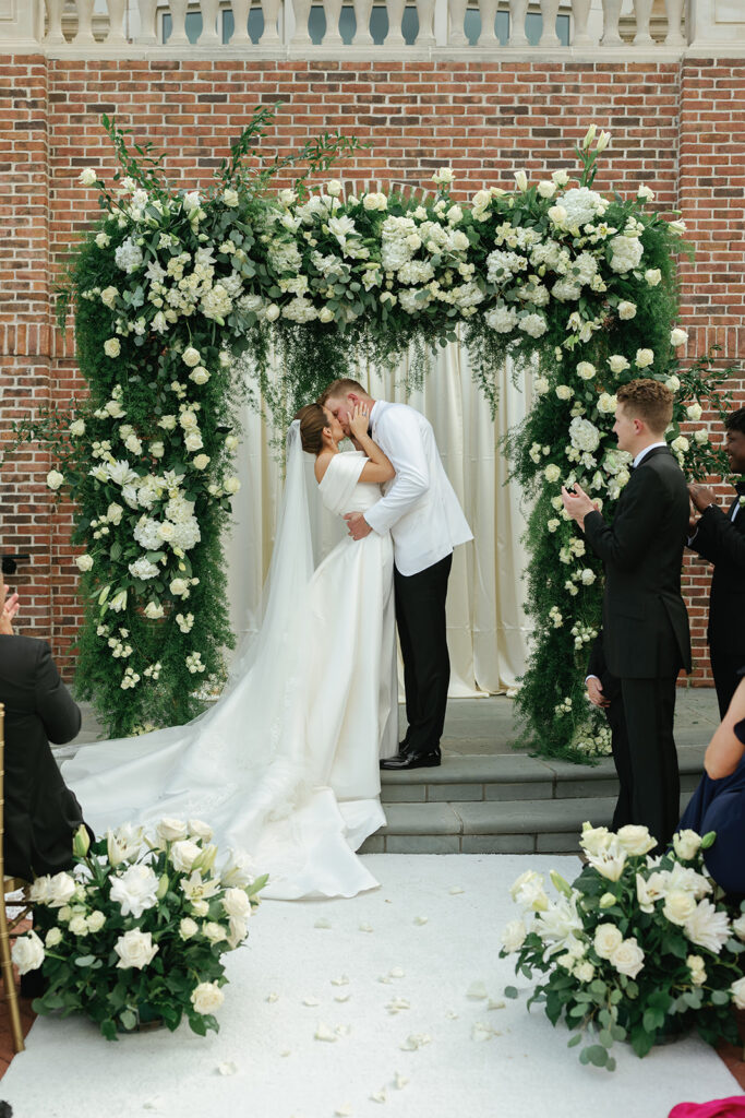 Bride and groom sharing their first kiss during a classic Texas country club wedding ceremony beneath an elegant white and green floral arch